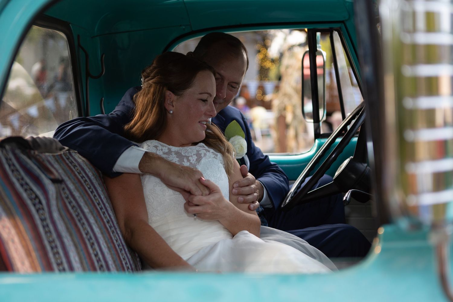 A bride in white dress sits in a vintage turquoise truck while holding a small pet.