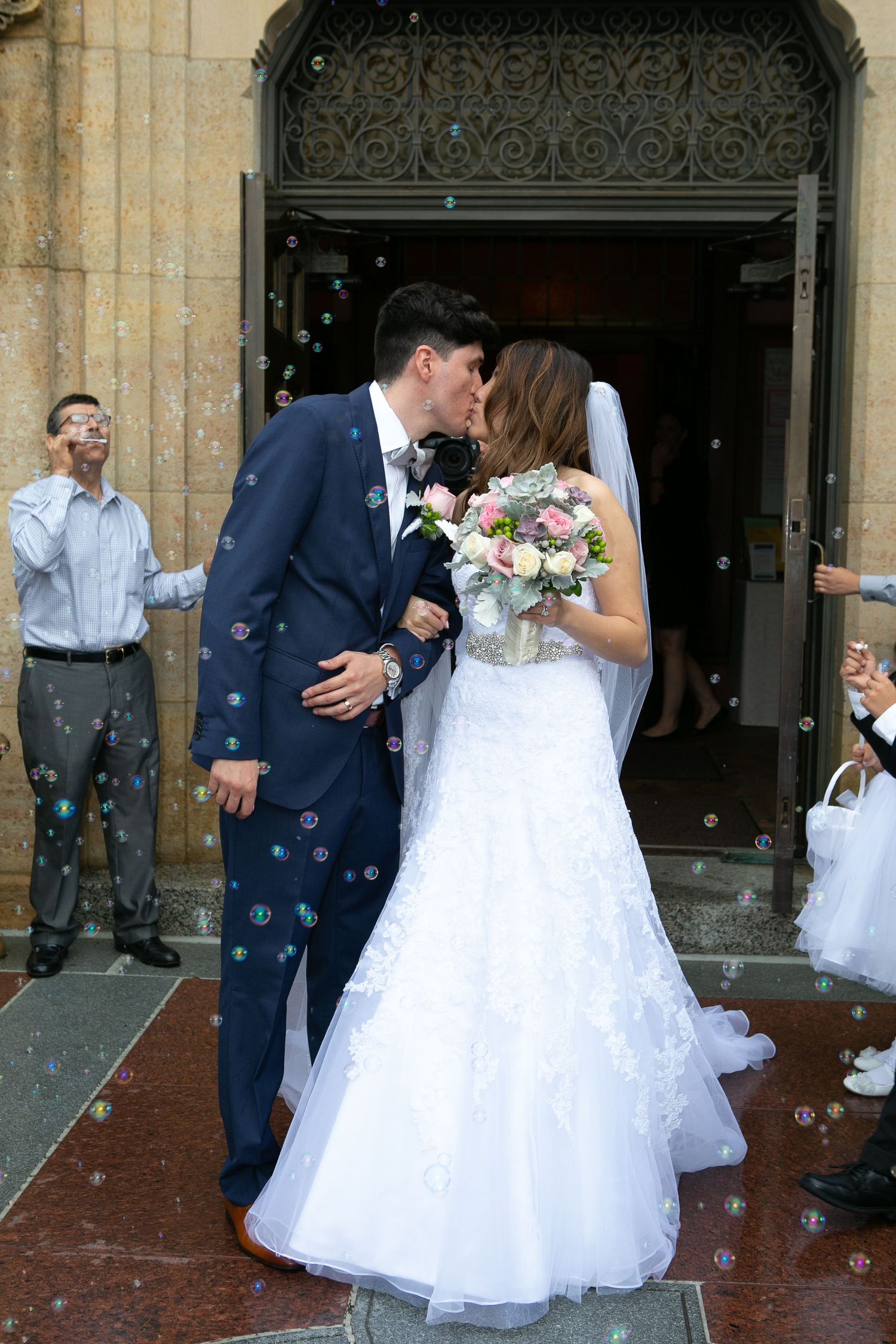 Newlyweds share first kiss under ornate church entrance as guests blow bubbles.