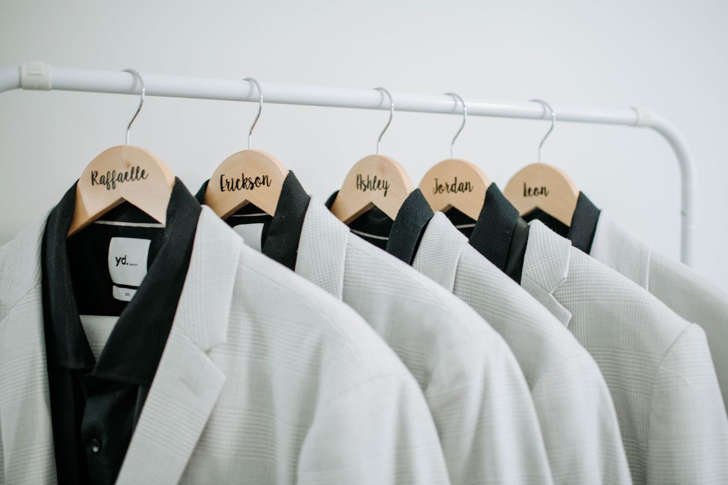 White suit jackets hanging on wooden hangers with groomsmen names labeled on each.