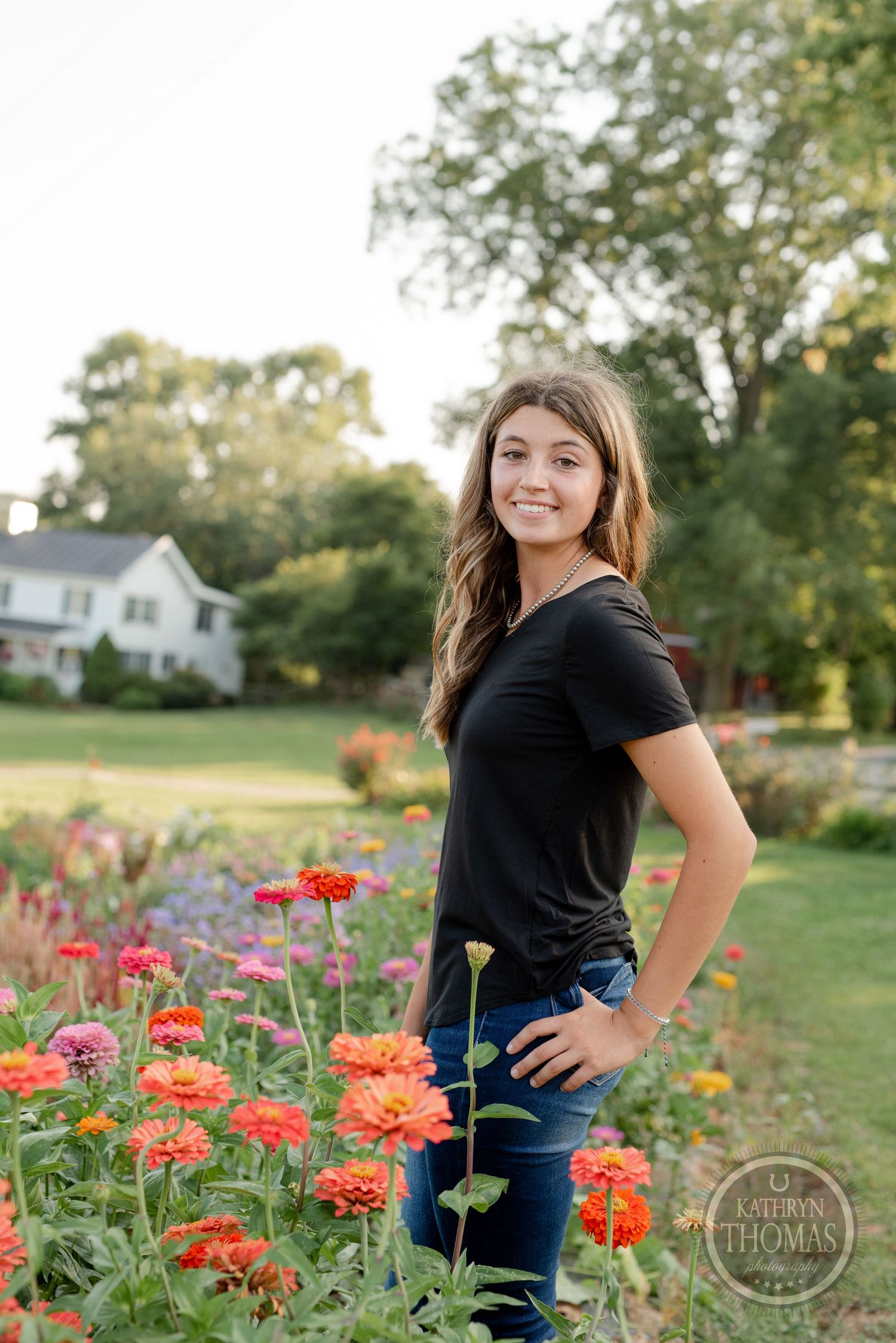 Senior portrait in a garden with vibrant orange and pink zinnias and a white farmhouse in the background.