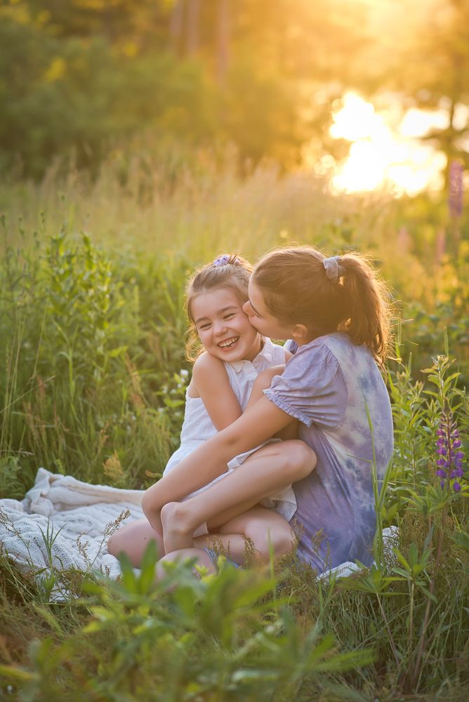two sisters hugging and giving little sister kiss on the cheek in beautiful wild flower field at sunset