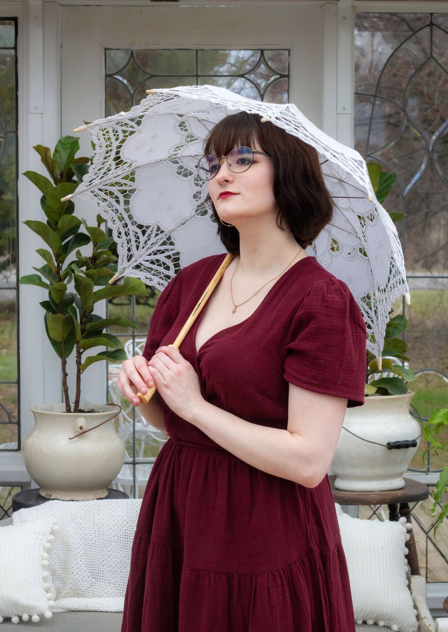 Model in burgundy dress poses with white lace parasol in front of greenhouse with potted plants.