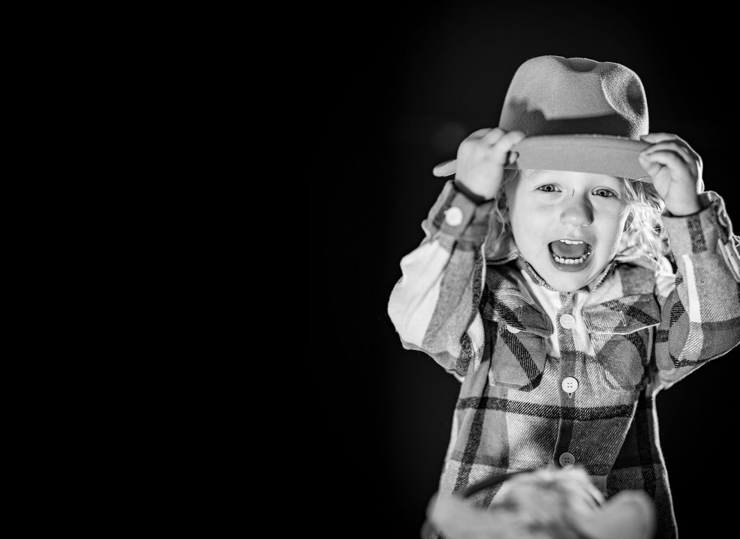 Excited young child wearing cowboy hat and plaid shirt laughing against black background in black and white photo.