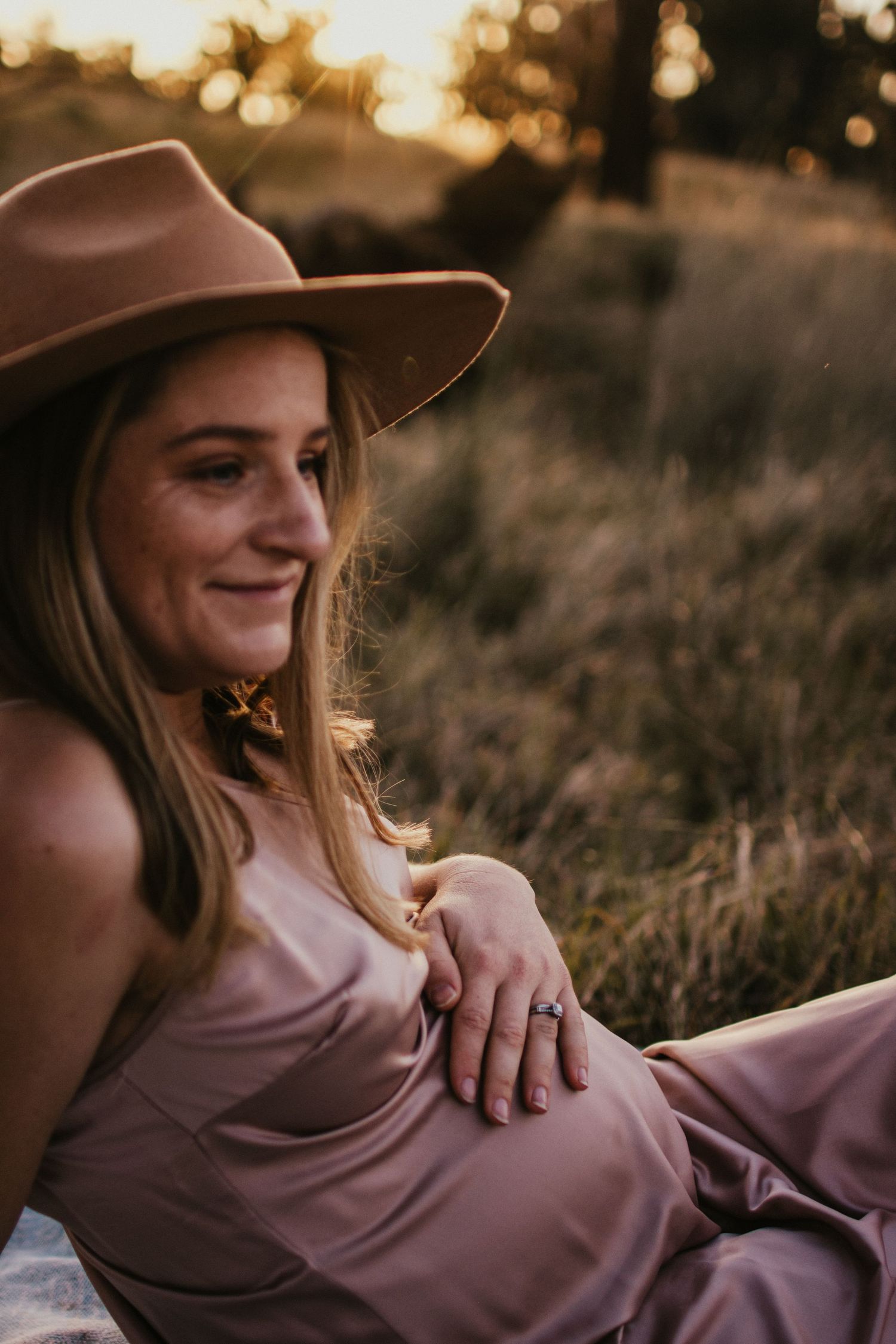 Pregnant mum in a bronze satin dress in a paddock at golden hour with her hand on her pregnant belly.
