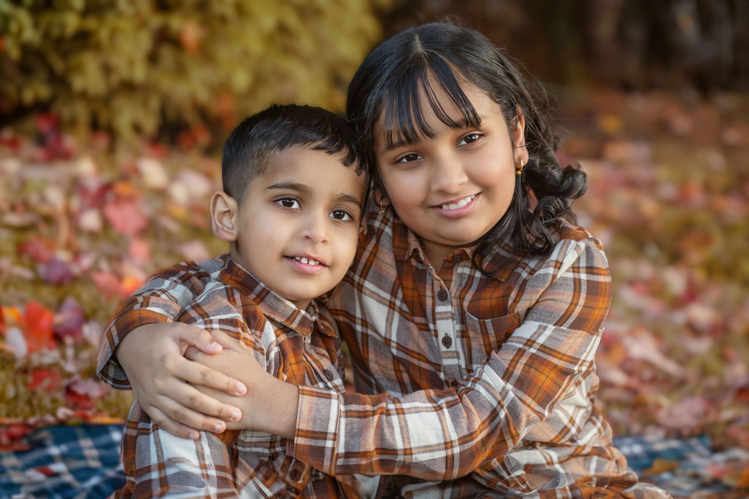 Two children in matching plaid shirts embrace while sitting on autumn leaves.