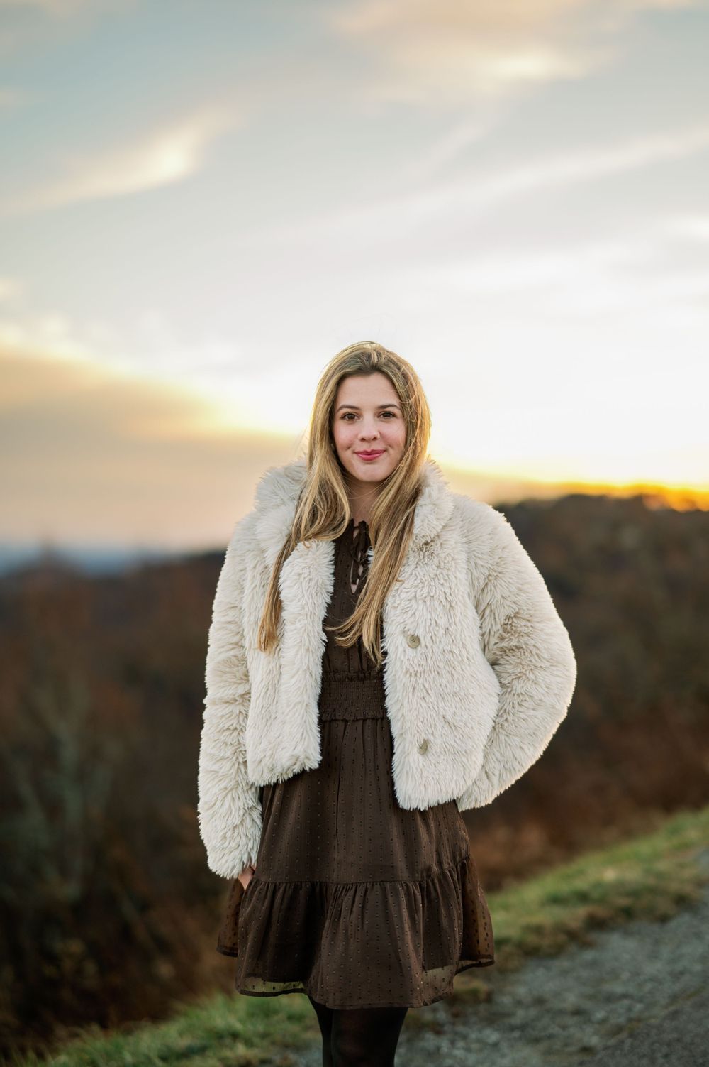 Portrait of person in fluffy white coat and brown dress against sunset mountain backdrop.