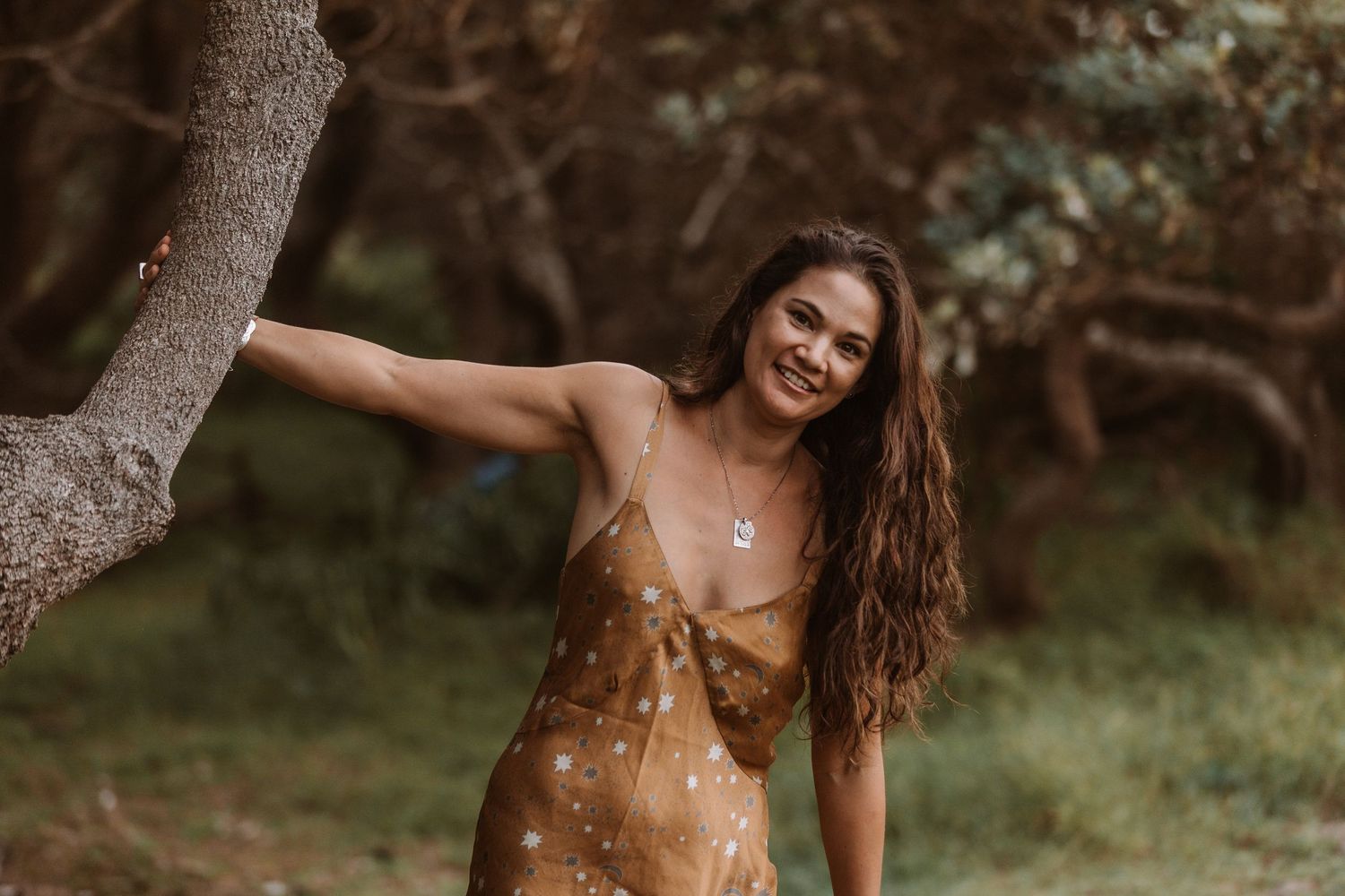 A sun-dappled portrait in nature shows a smiling figure in a brown floral dress leaning against a tree trunk. Portrait photography.