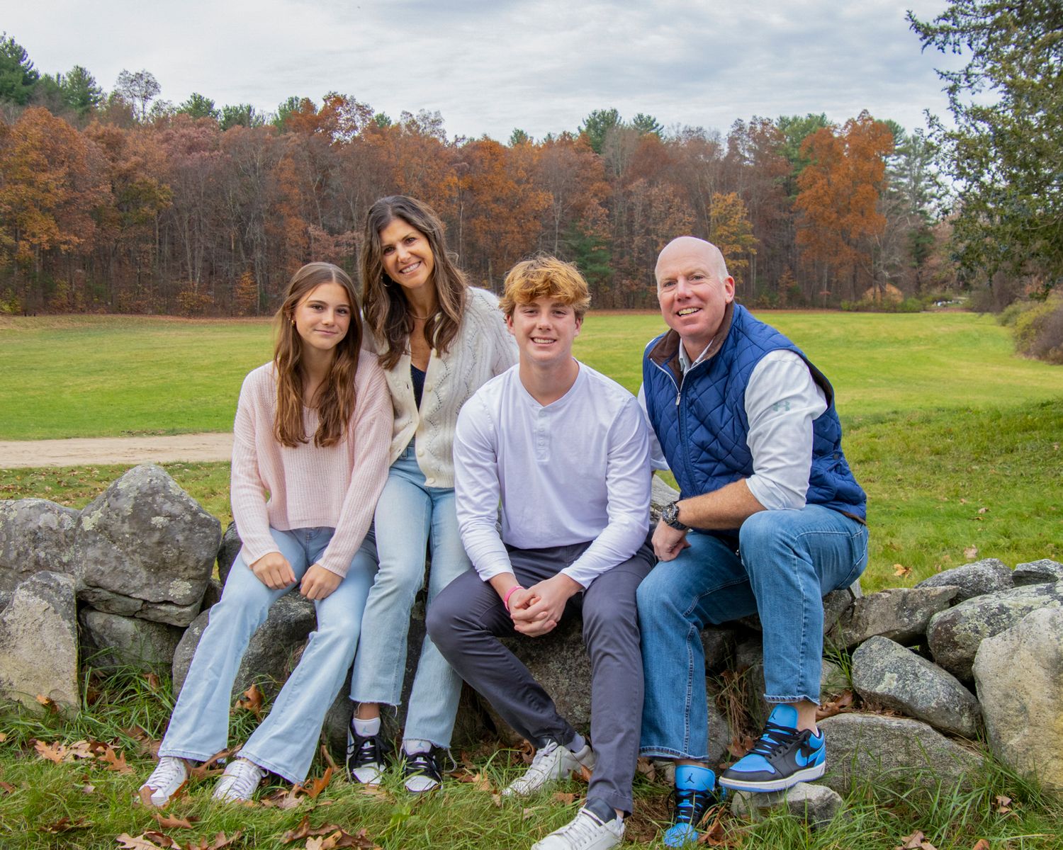 A family sits together on a stone wall surrounded by autumn foliage in a park setting.