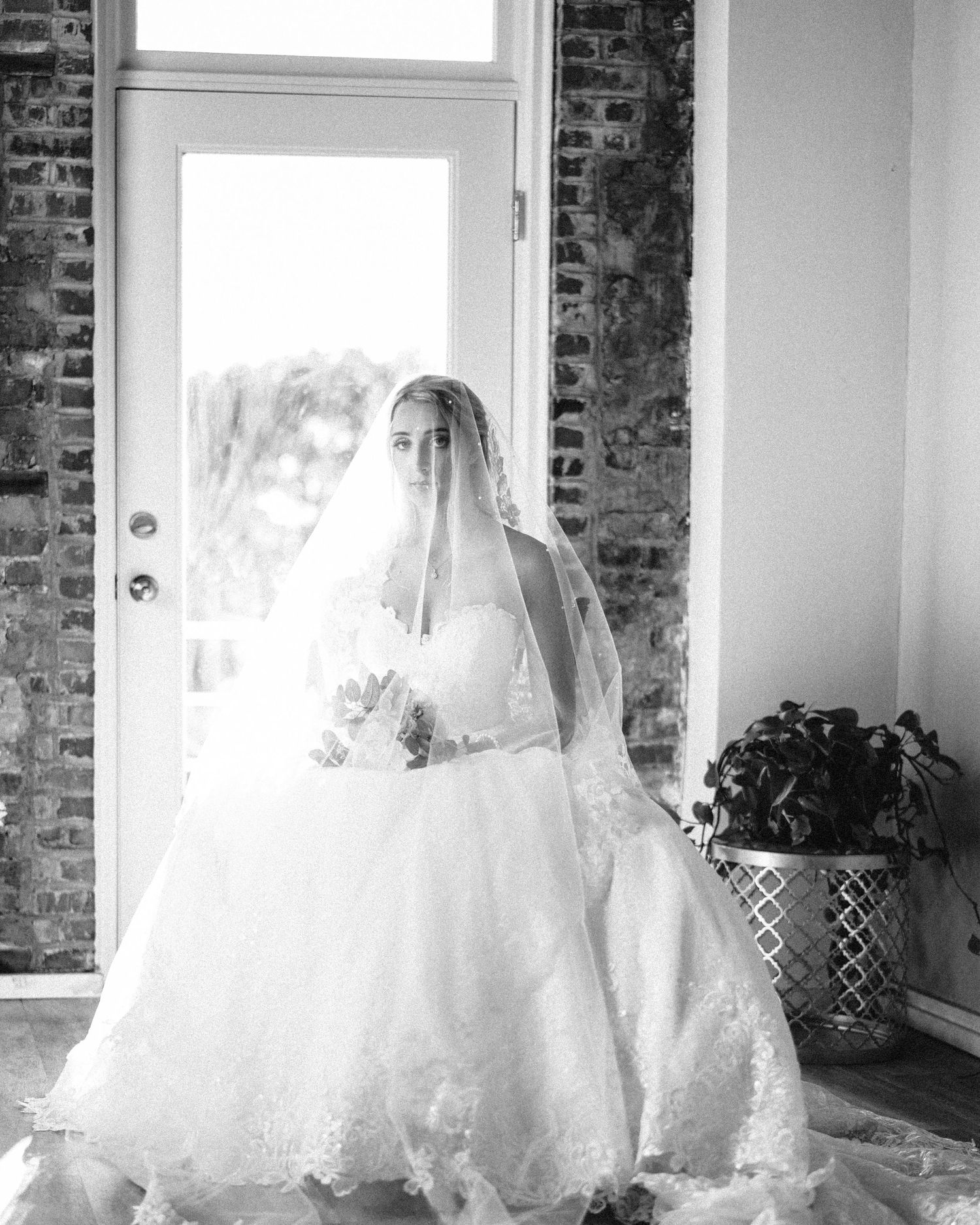 A seated bride in a full white dress and veil poses near a window with exposed brick walls.