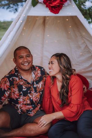 A couple shares intimate moments while sitting in front of a white teepee tent decorated with red flowers.