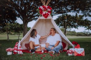Couple lounging together near a decorated white teepee tent with red floral accents at sunset.