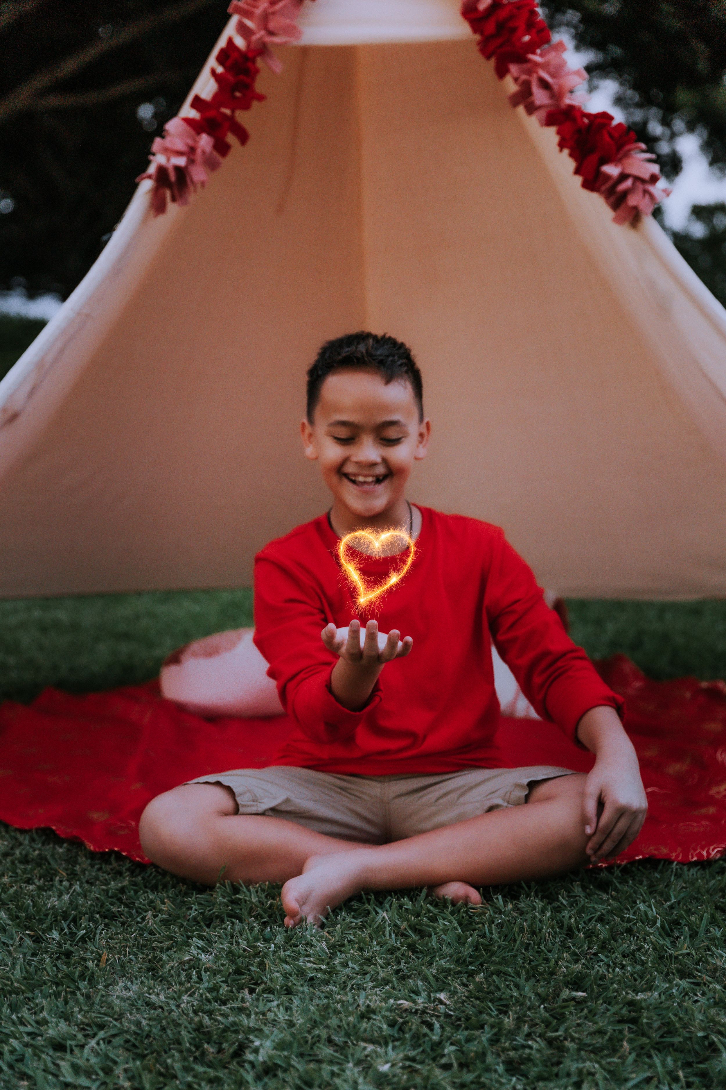Child in red clothing sits on grass reading near a white tent decorated with pink flowers.