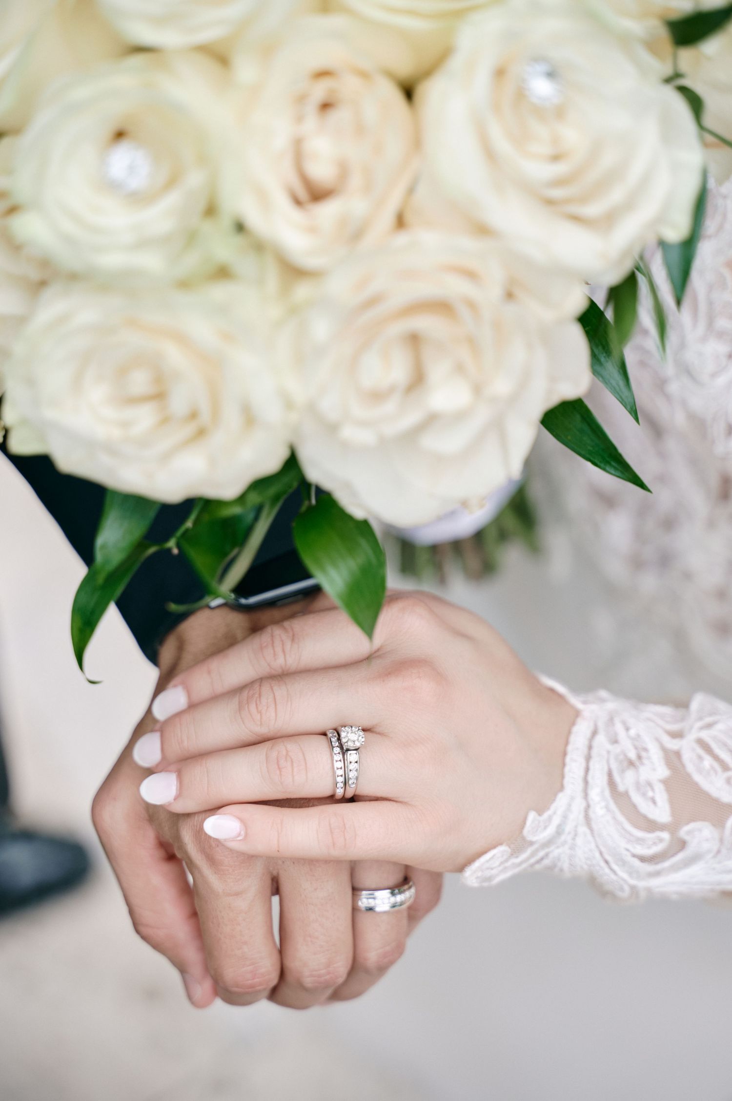 Close-up of hands holding white roses showing wedding rings and lace sleeve detail.