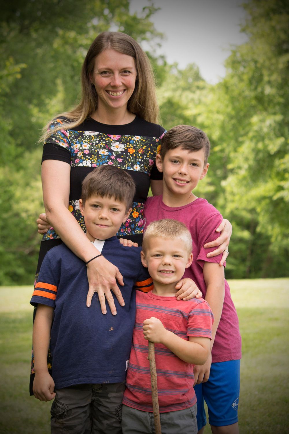 A smiling mother poses outdoors with her three young sons in a lush green park setting.