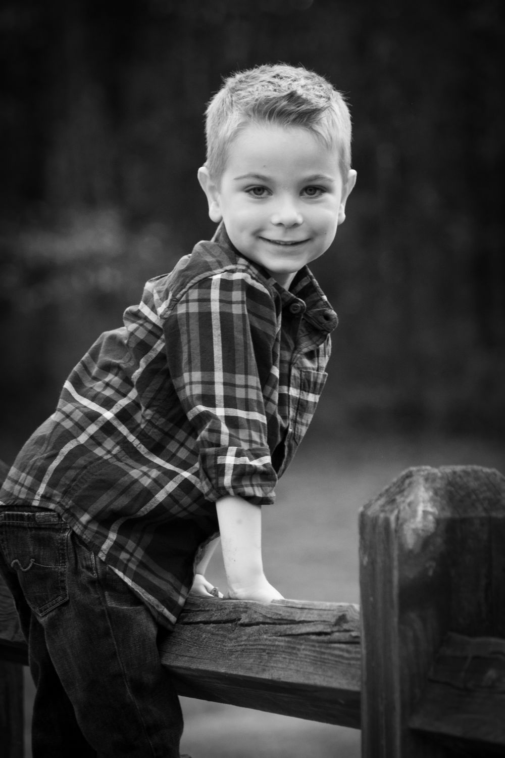 Adorable young boy in plaid shirt leans on wooden fence smiling in black and white photo.