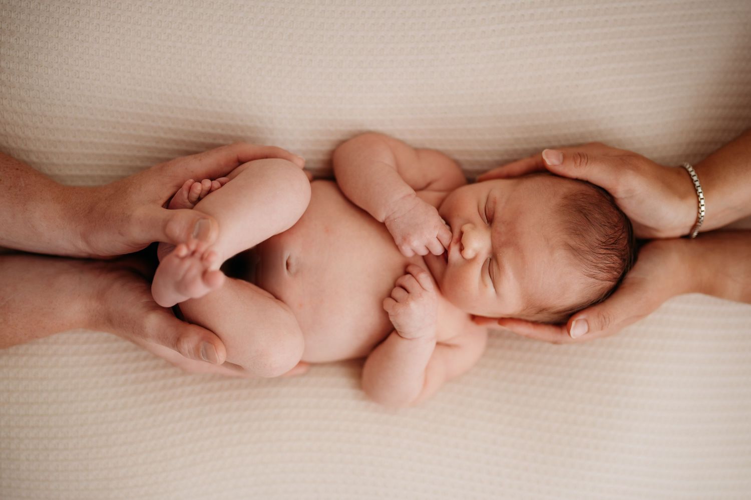 Sleeping newborn cradled between adult hands on beige background.