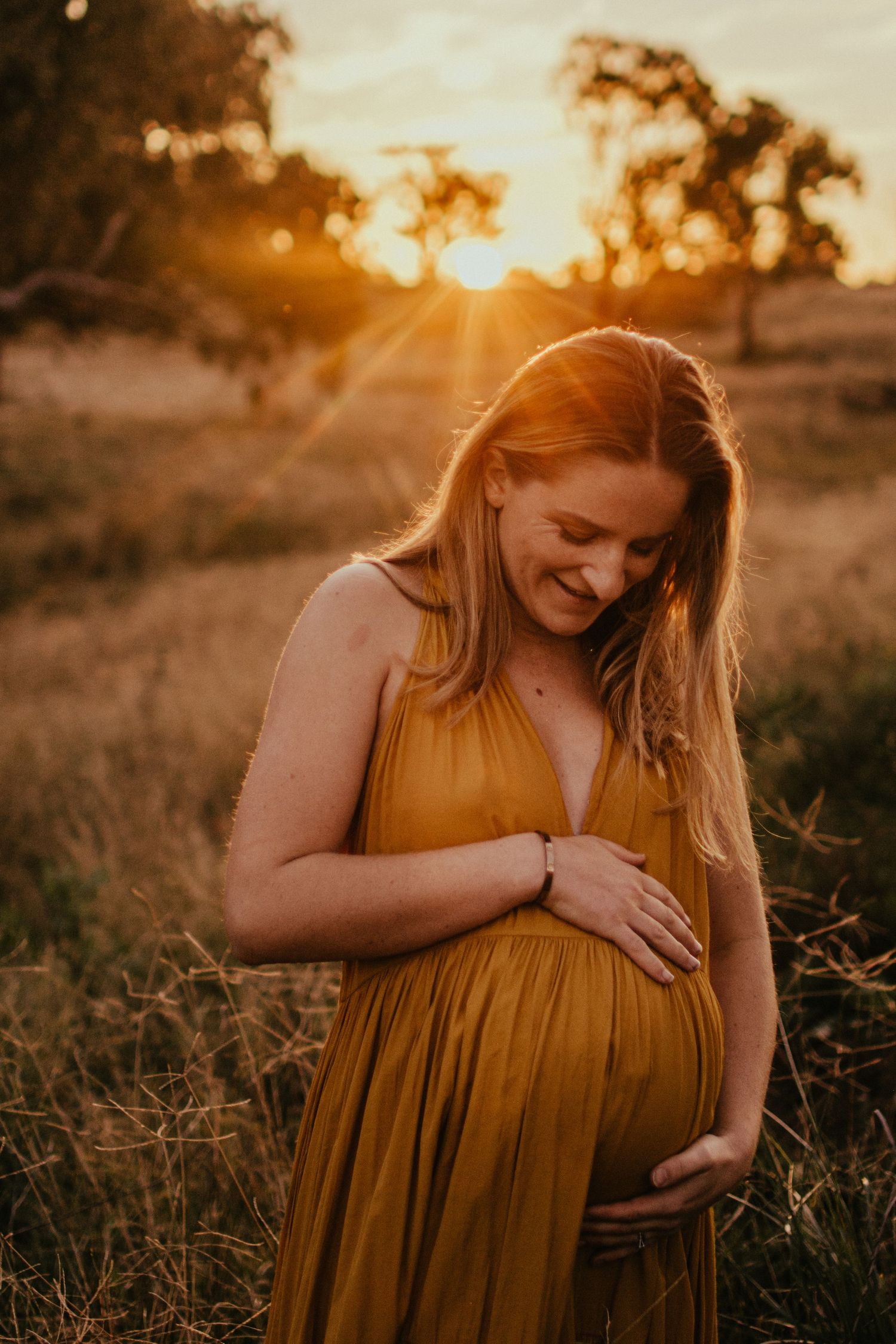 Pregnant Mum, in a golden dress standing in a long grass paddock with a golden sunset. Looking down at her growing belly