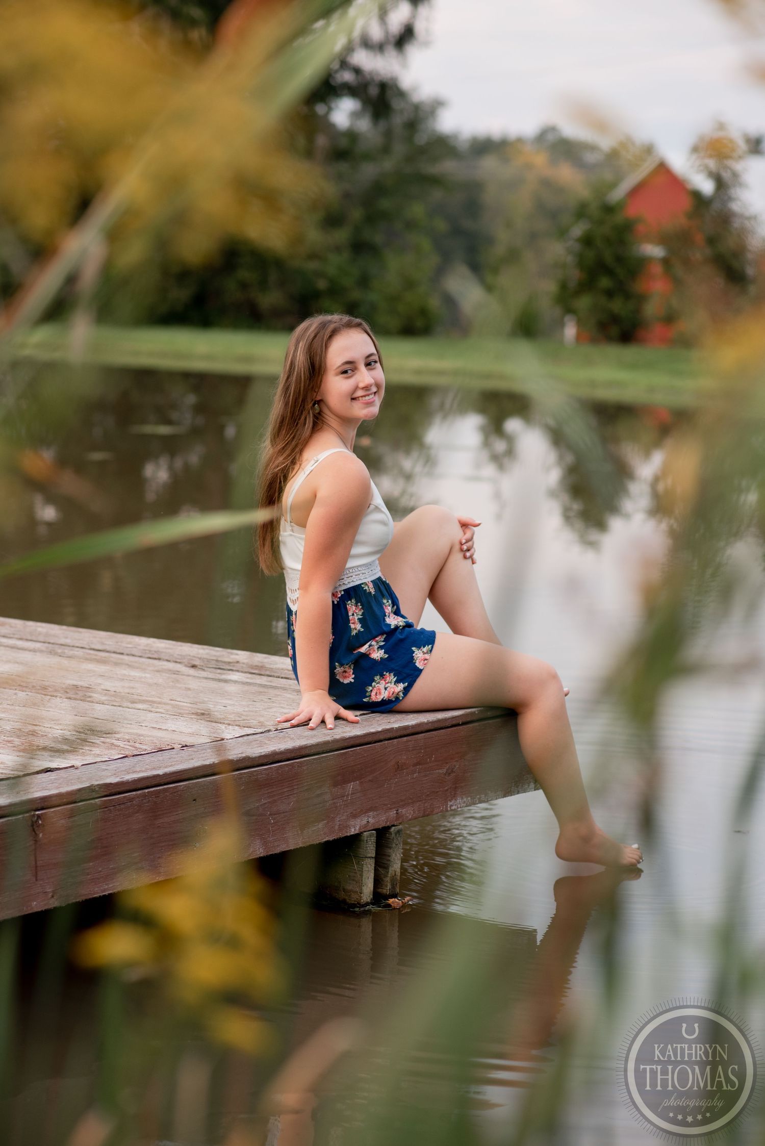 Outdoor portrait at a pond with someone sitting on wooden dock wearing navy floral shorts and white tank top.