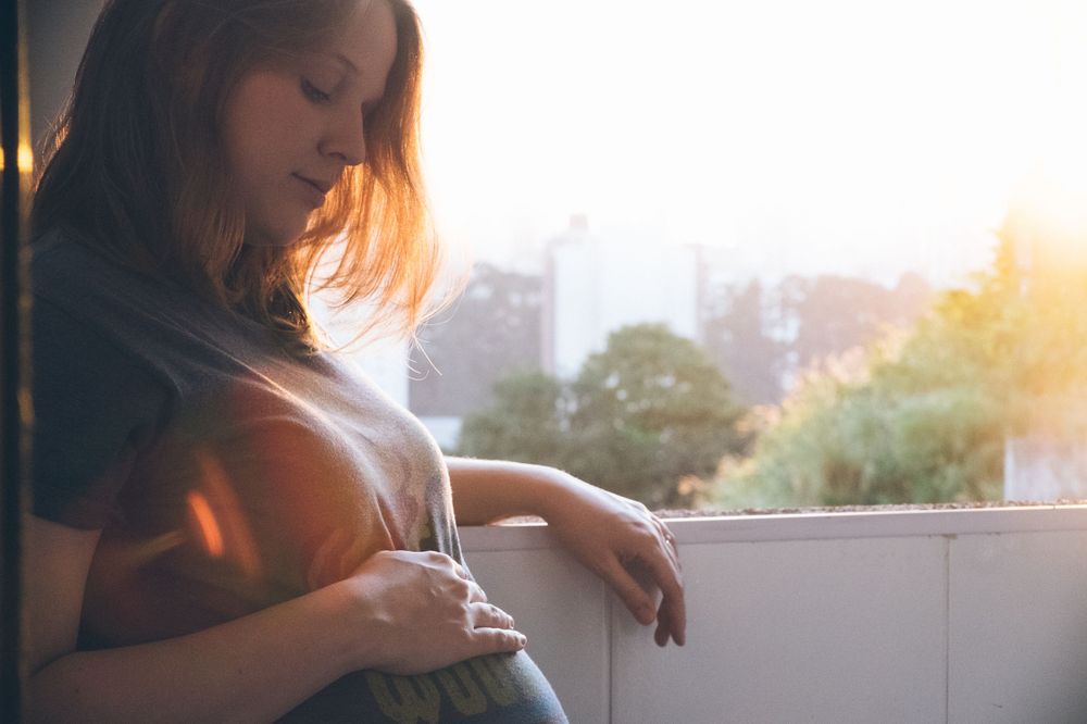 Side view of pregnant person standing by window during sunset.