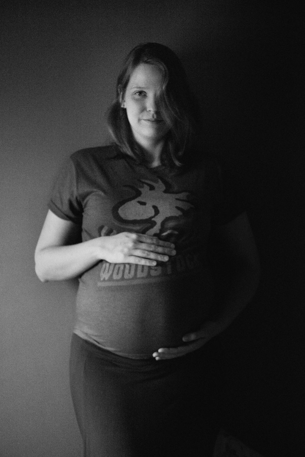 Black and white portrait of pregnant person wearing graphic t-shirt against dark background.