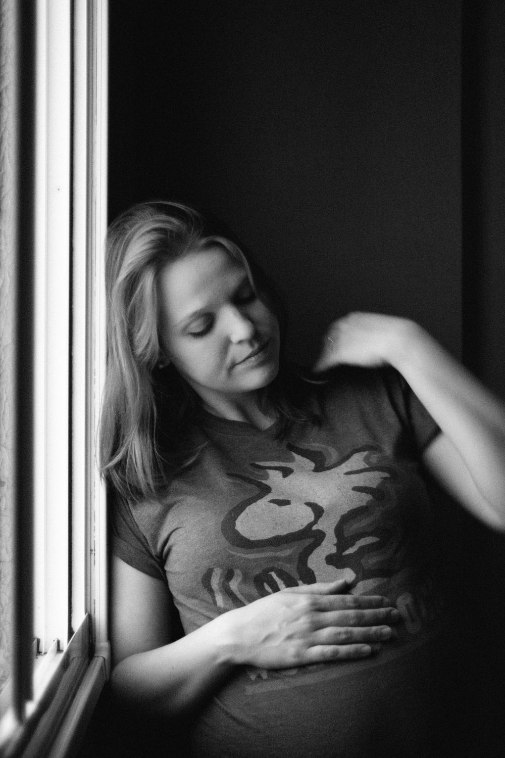 Black and white window portrait of pregnant person in graphic t-shirt with gentle expression.
