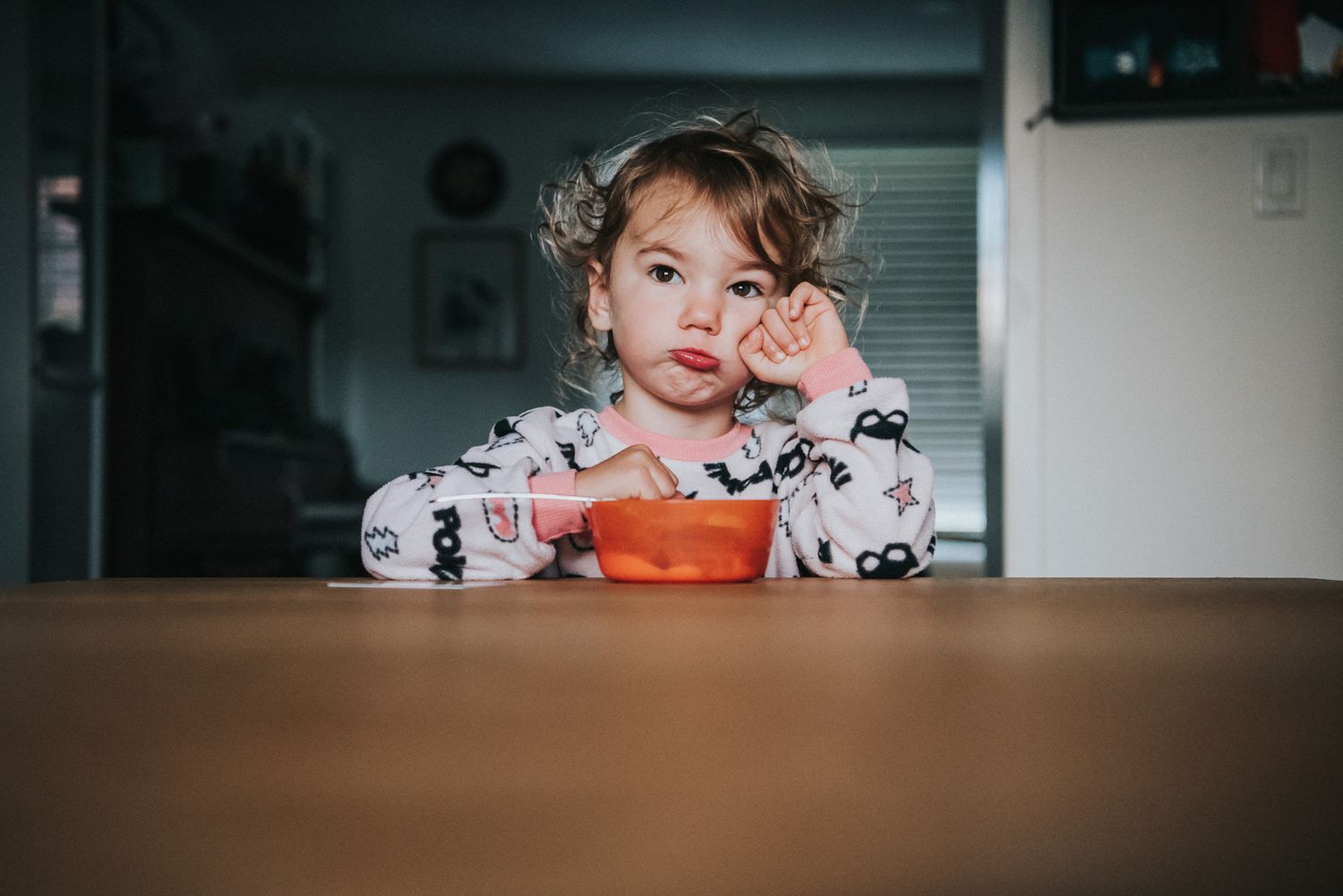 A young child sits at a table with an orange bowl a small pout on her face as she rests her hand on cheek.