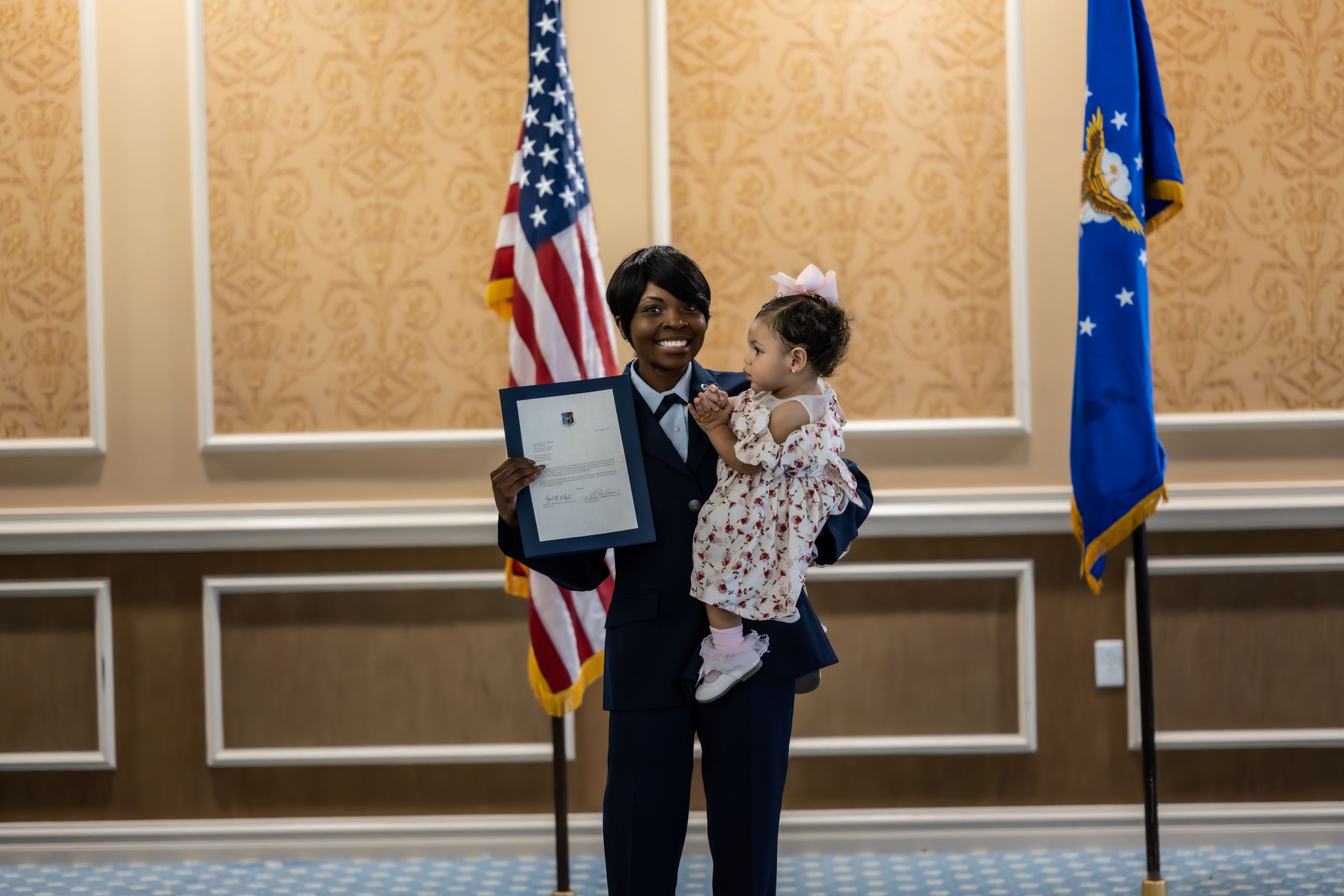 Military ceremony participant displays official document while holding child near flags.