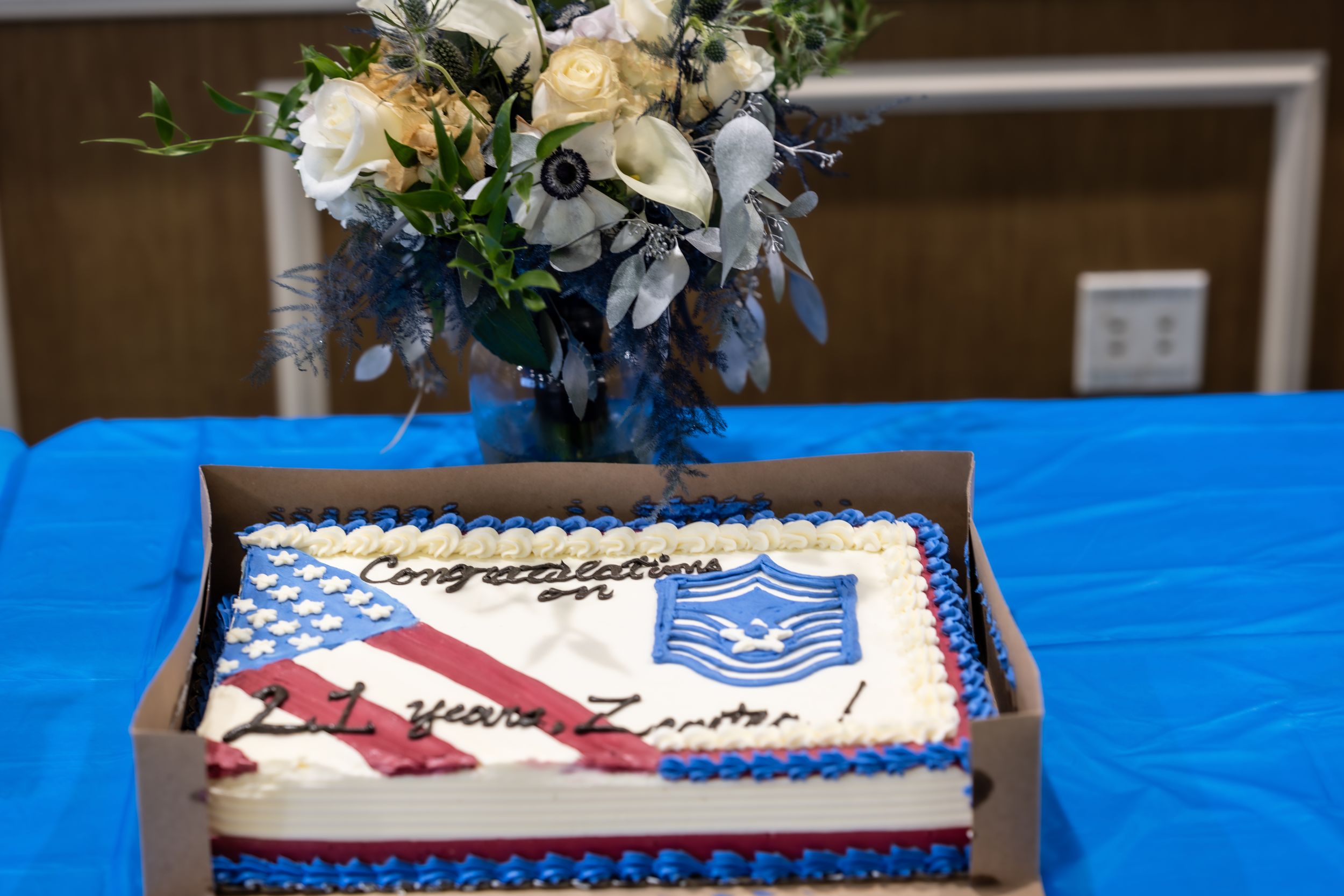 Decorated cake with Air Force emblem sits on blue tablecloth with floral arrangement.