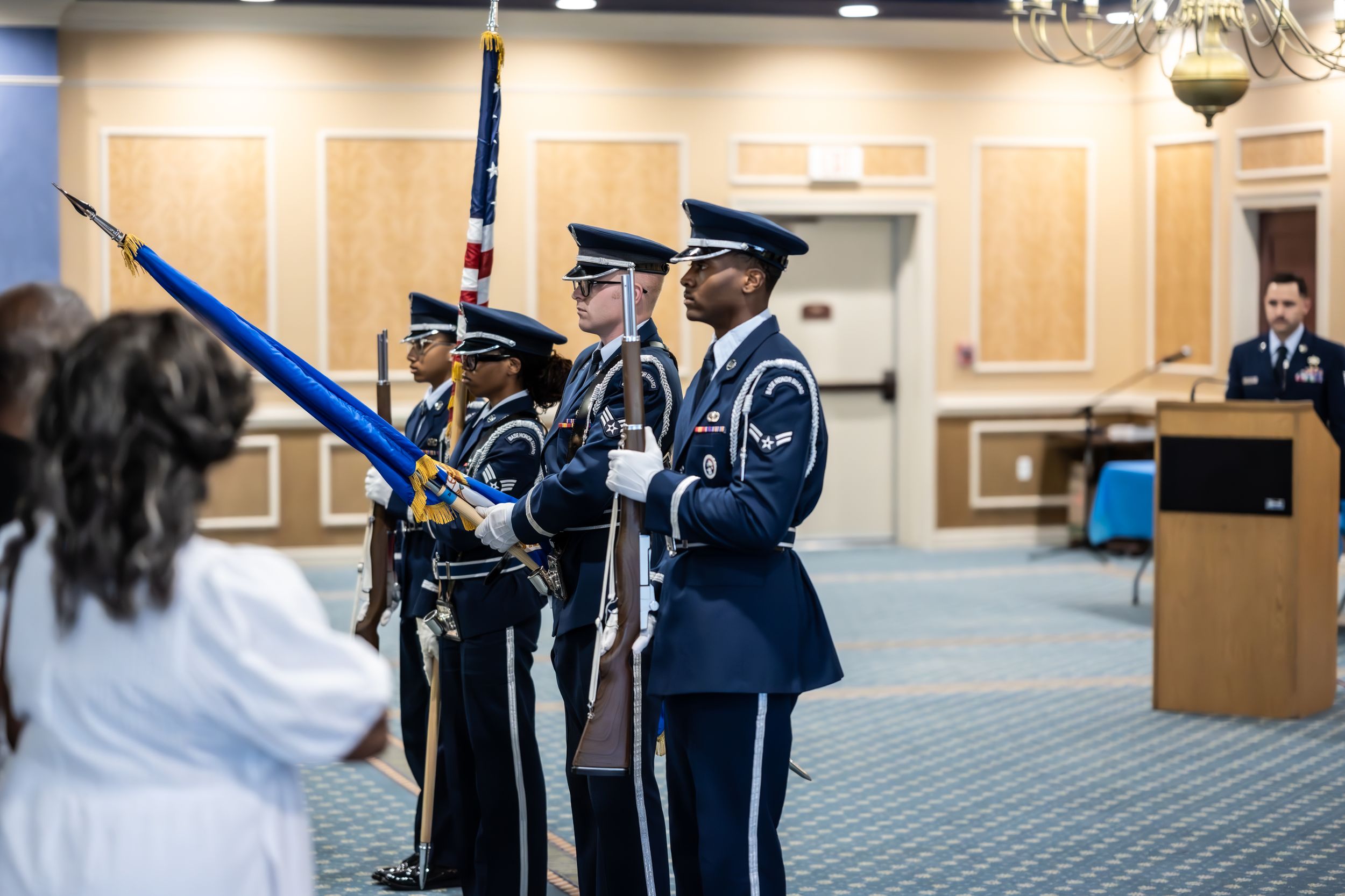 Military personnel stand in formation during a ceremony in an elegant carpeted room with beige walls.