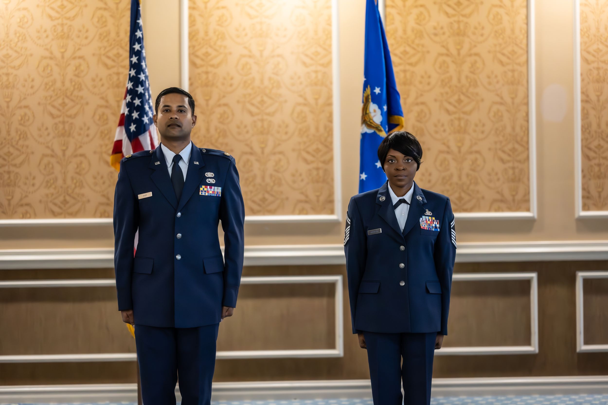 Military officers stand formally during an official ceremony with flags and decorative wall panels.
