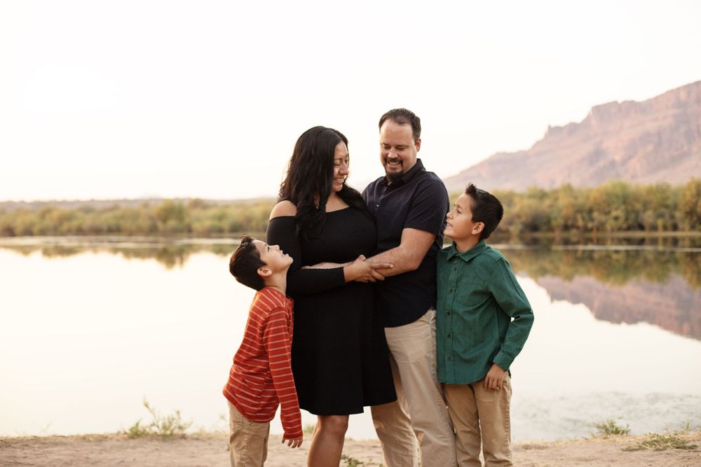Family members embrace while sitting on rocks near a desert lake with mountains and autumn foliage.