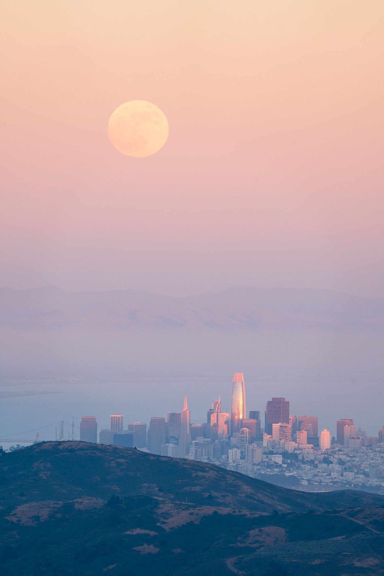 Full moon rises over San Francisco skyline at dusk with pink and purple skies casting ethereal glow over cityscape.