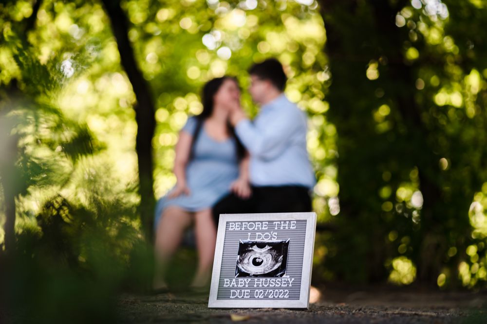Couple holds pregnancy announcement sign in sunlit wooded path.