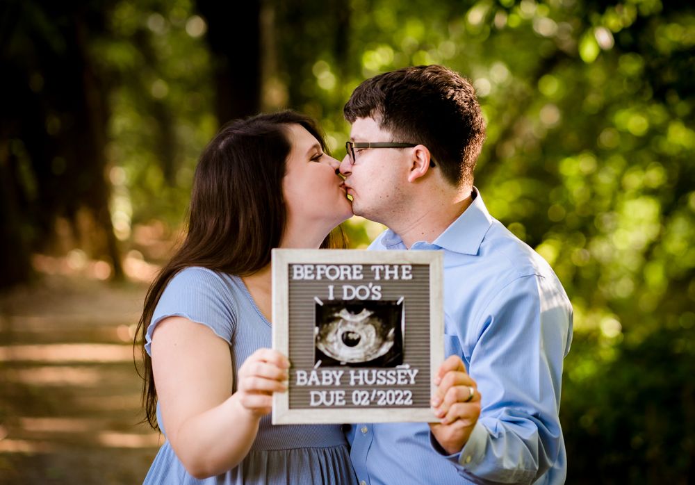 Couple shares intimate moment while holding baby announcement sign in park setting.