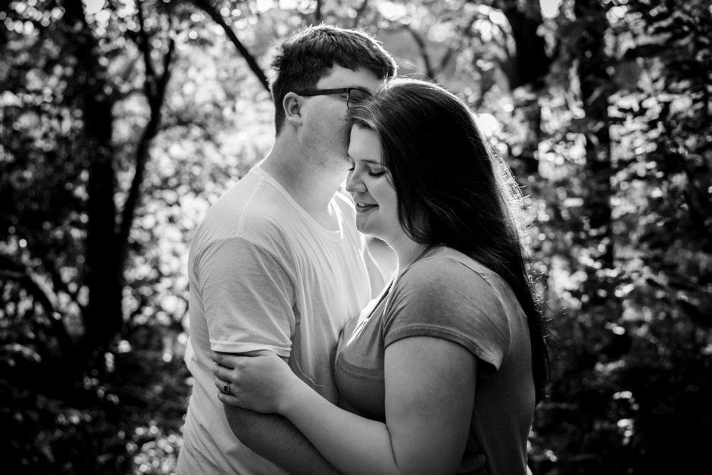 An intimate black and white portrait of a couple embracing in a forest setting.