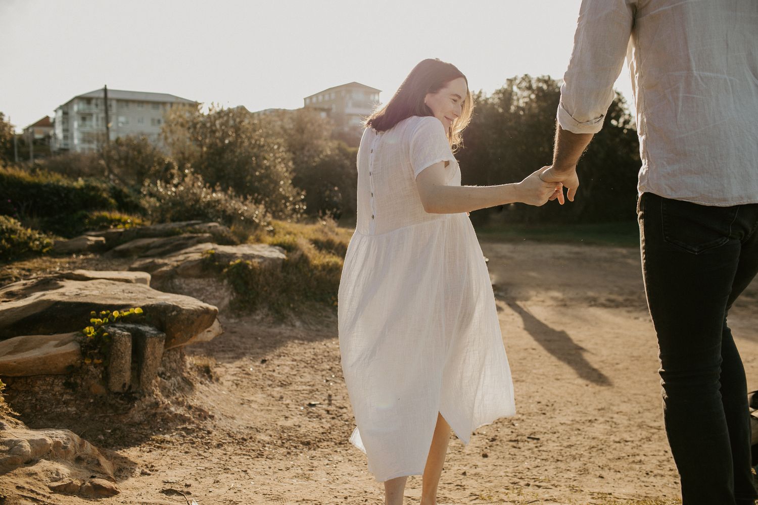 A woman in a white dress holds a mans hand while walking along a sunny outdoor path at golden hour.