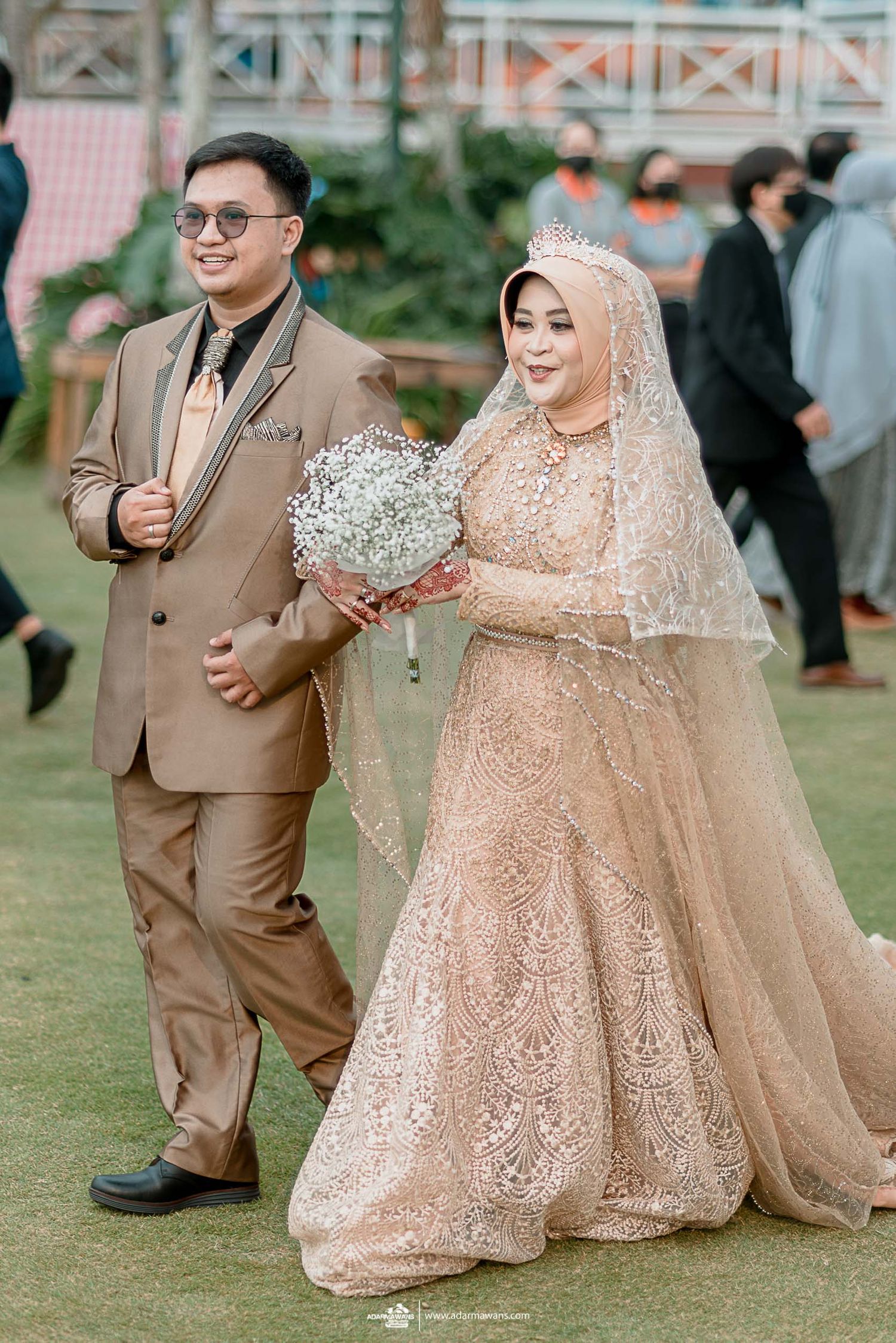 Couple walks together on grass holding baby's breath bouquet at outdoor wedding venue.