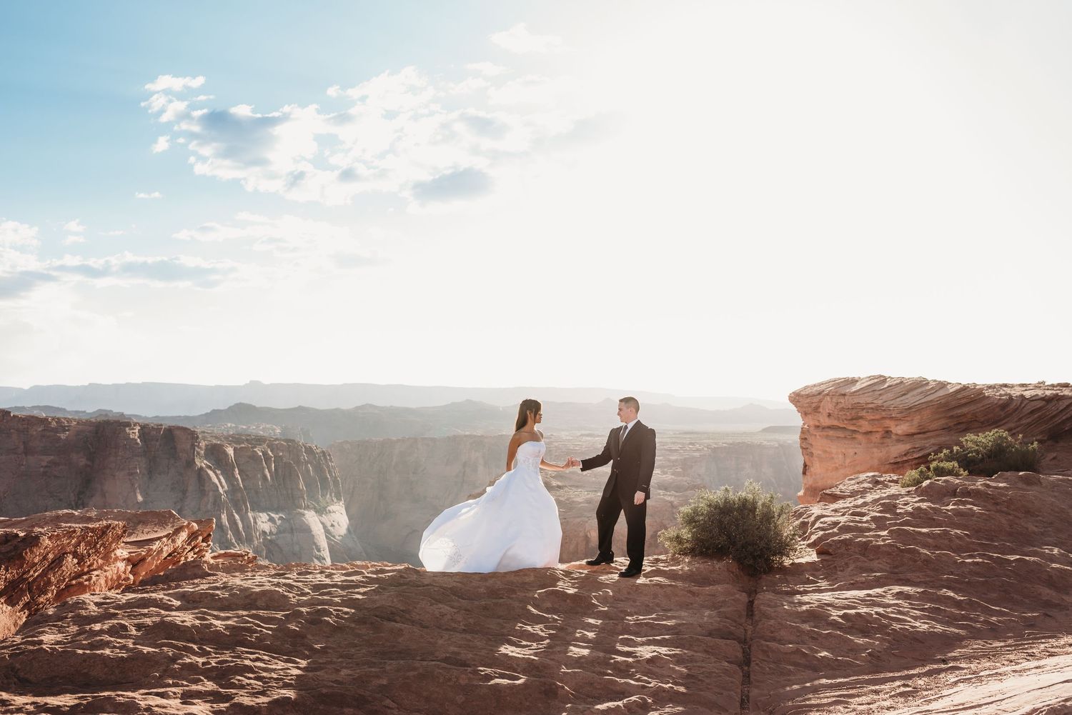 A couple in wedding attire stands on red rocks overlooking a scenic canyon vista.