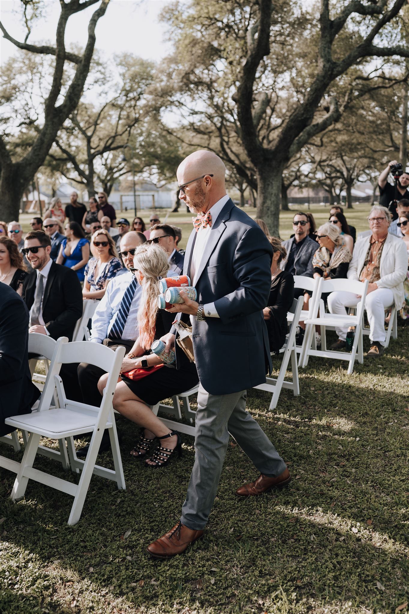 An elegantly dressed guest walks down the aisle at an outdoor wedding ceremony under oak trees.