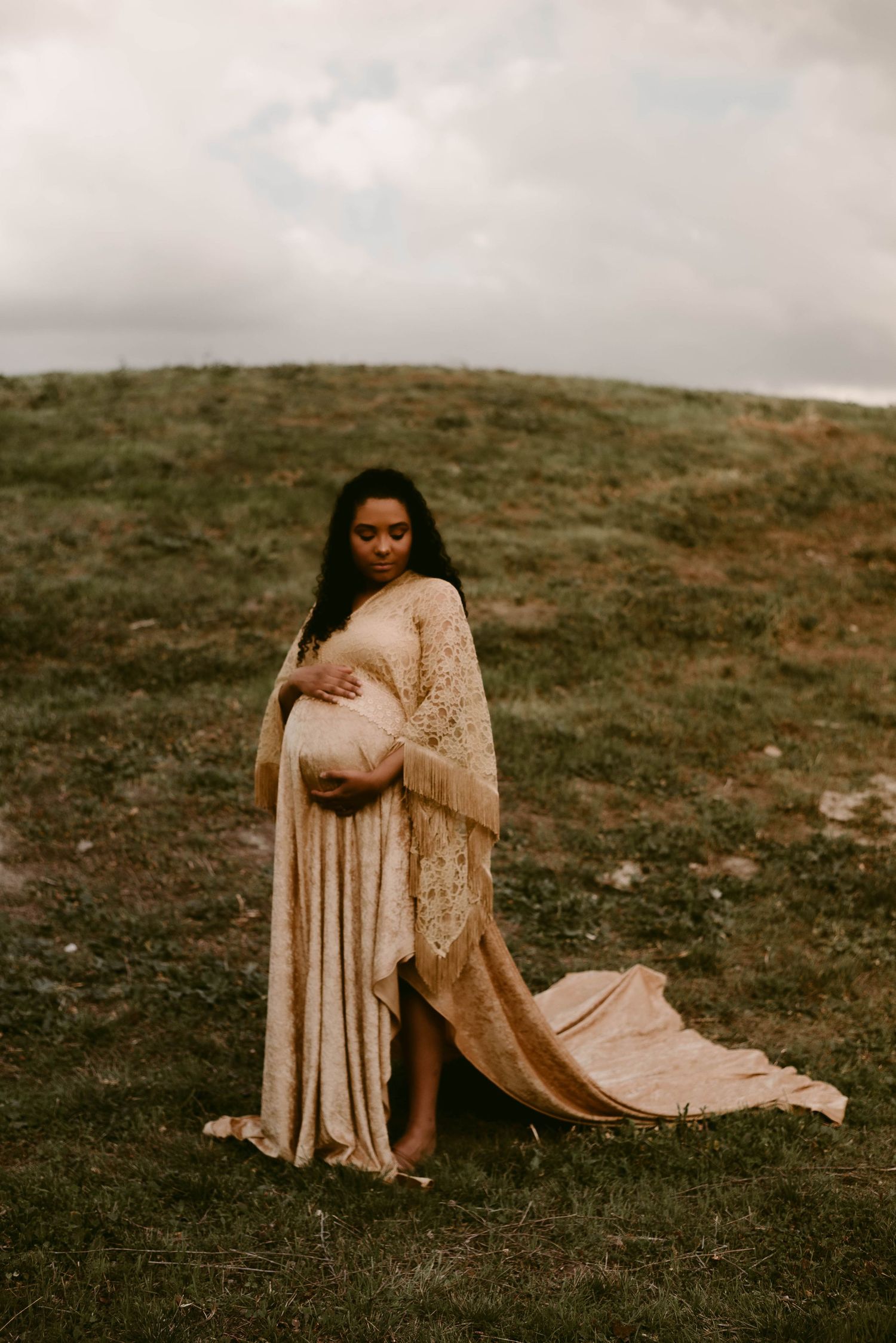Ethereal maternity portrait with long flowing dress and cape on grassy hilltop.