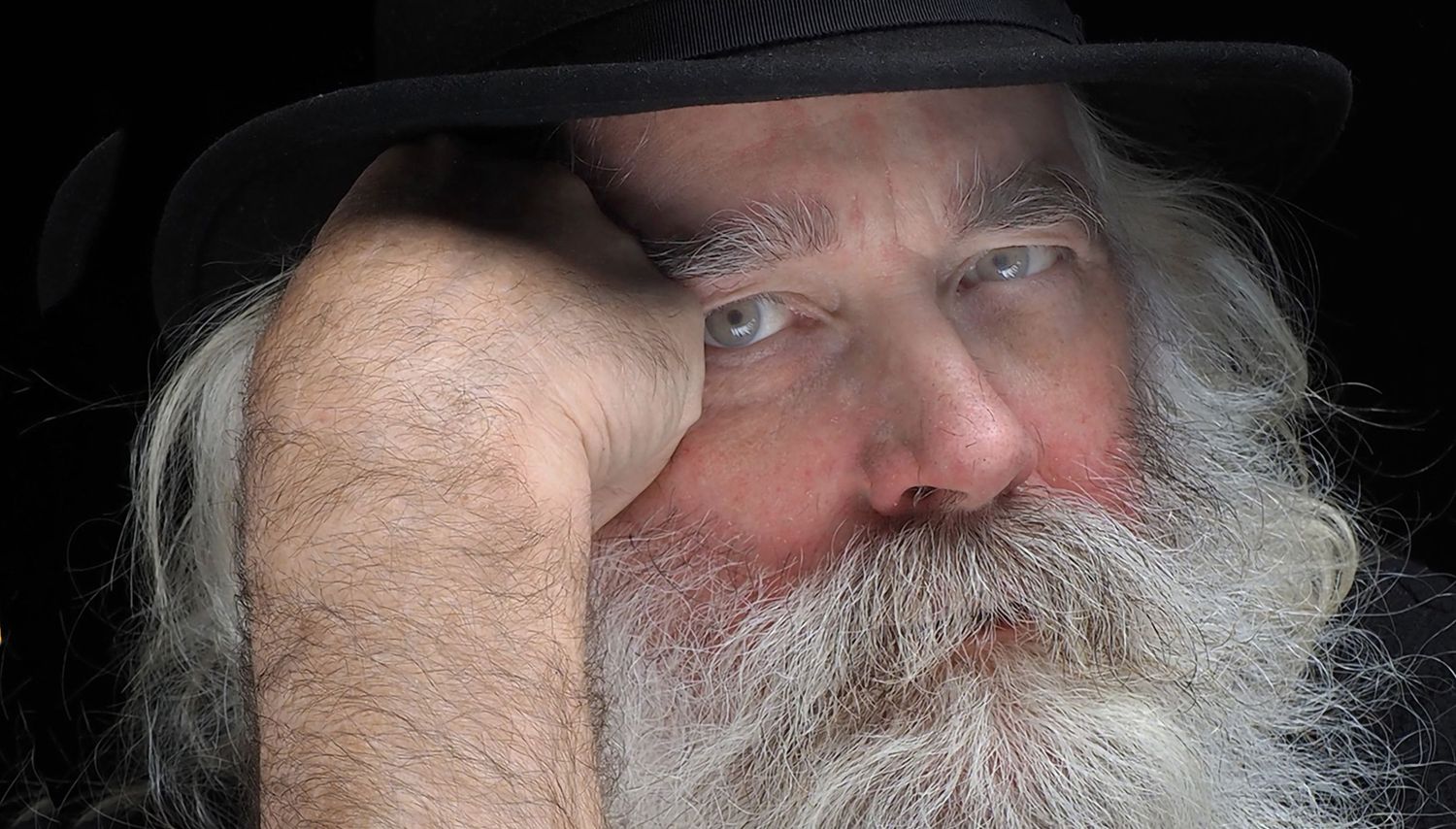 Close-up portrait of a person wearing a black hat with a white beard against dark background.