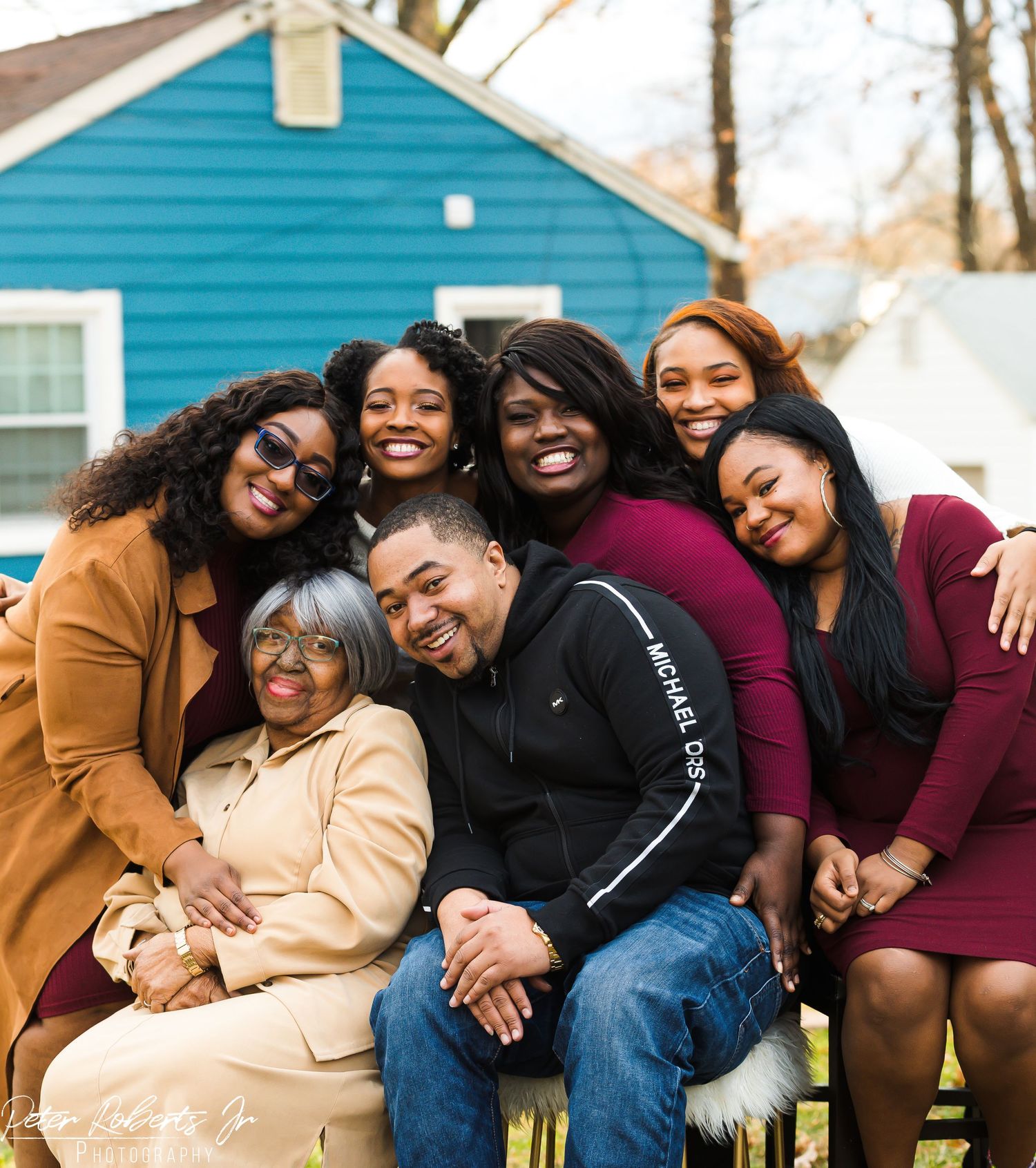Large joyful family group of seven women and one man posing together outdoors near a blue house.