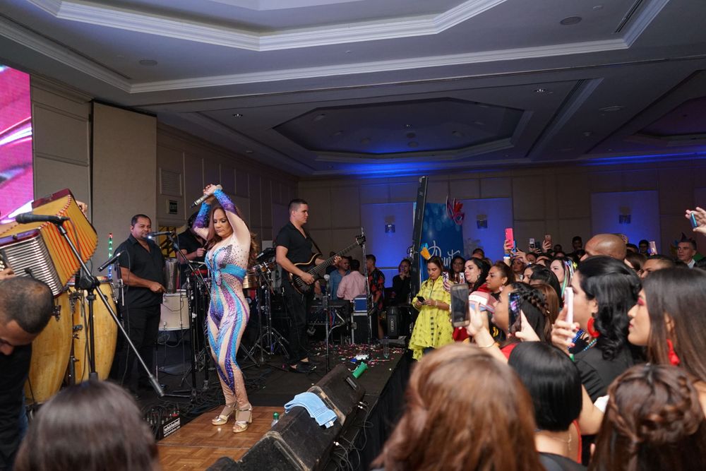 Latin band performs on stage at an energetic indoor concert event with a large dancing crowd.