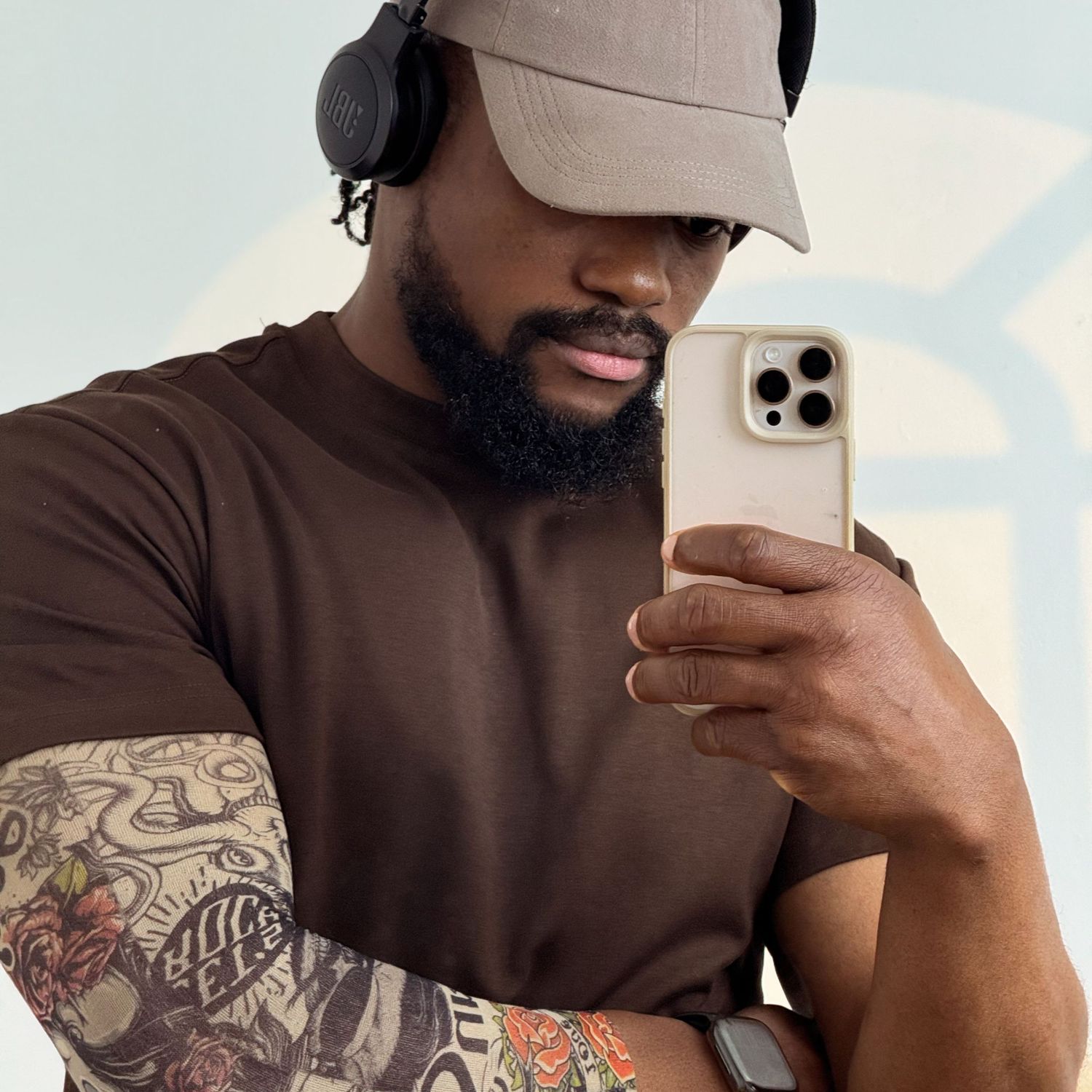 Tattooed man in brown tee and beige cap with headphones takes a mirror selfie with iPhone.