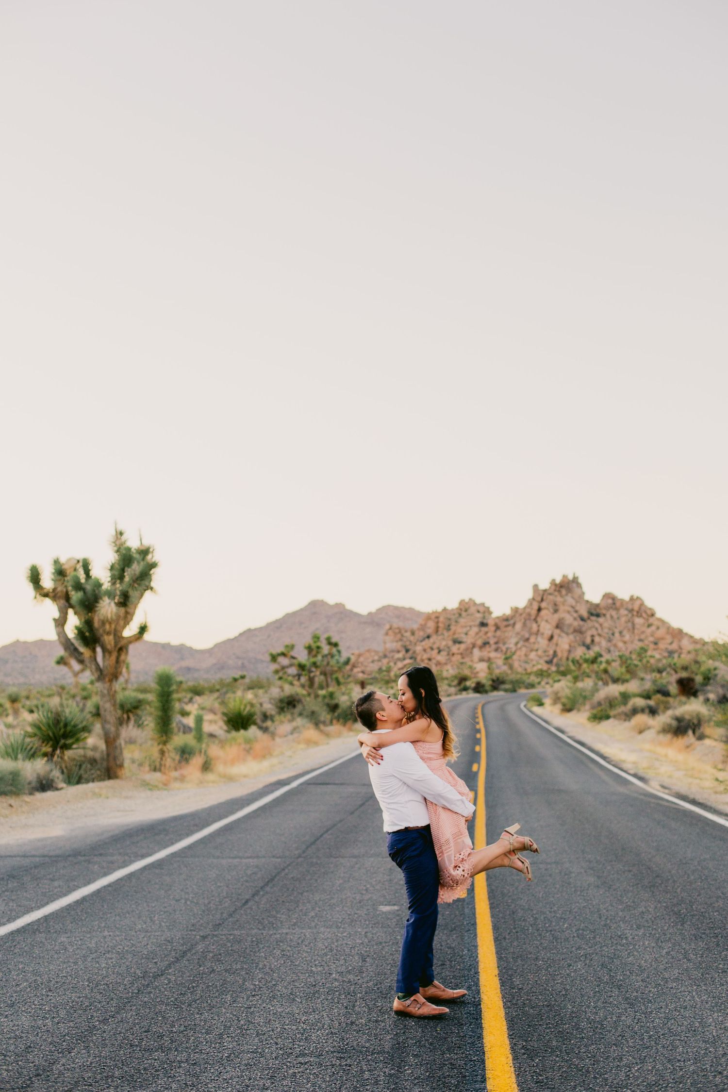 Couple embracing on an empty desert road with Joshua trees and mountains in the background.