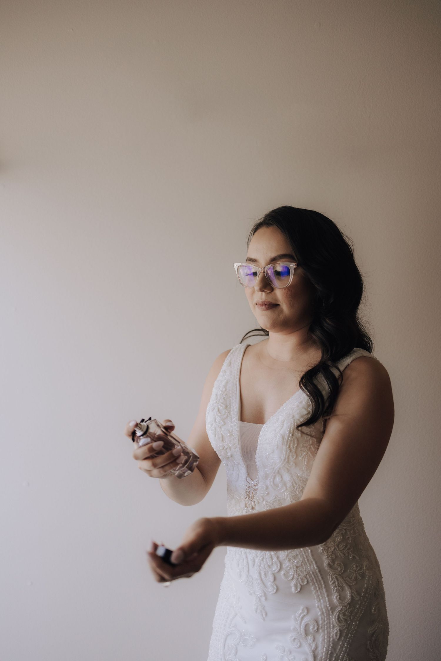 A person in a lace wedding dress and glasses stands against a neutral wall.
