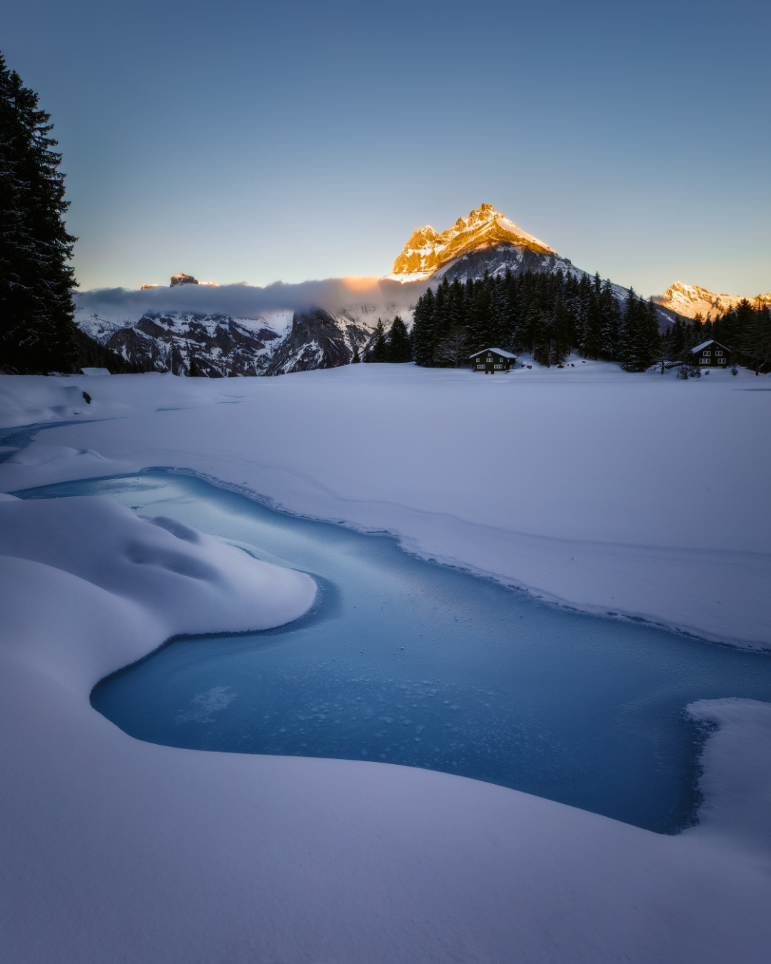 Mountain peak catches golden sunlight above a curved frozen lake surrounded by deep snow.