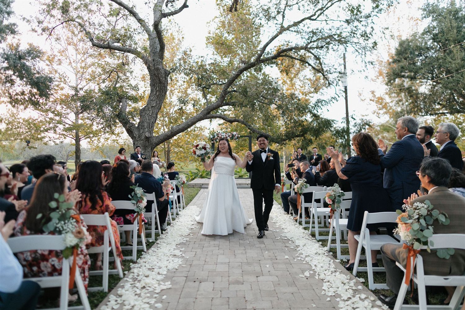 Wedding recessional along stone pathway with guests seated on white chairs under large trees.