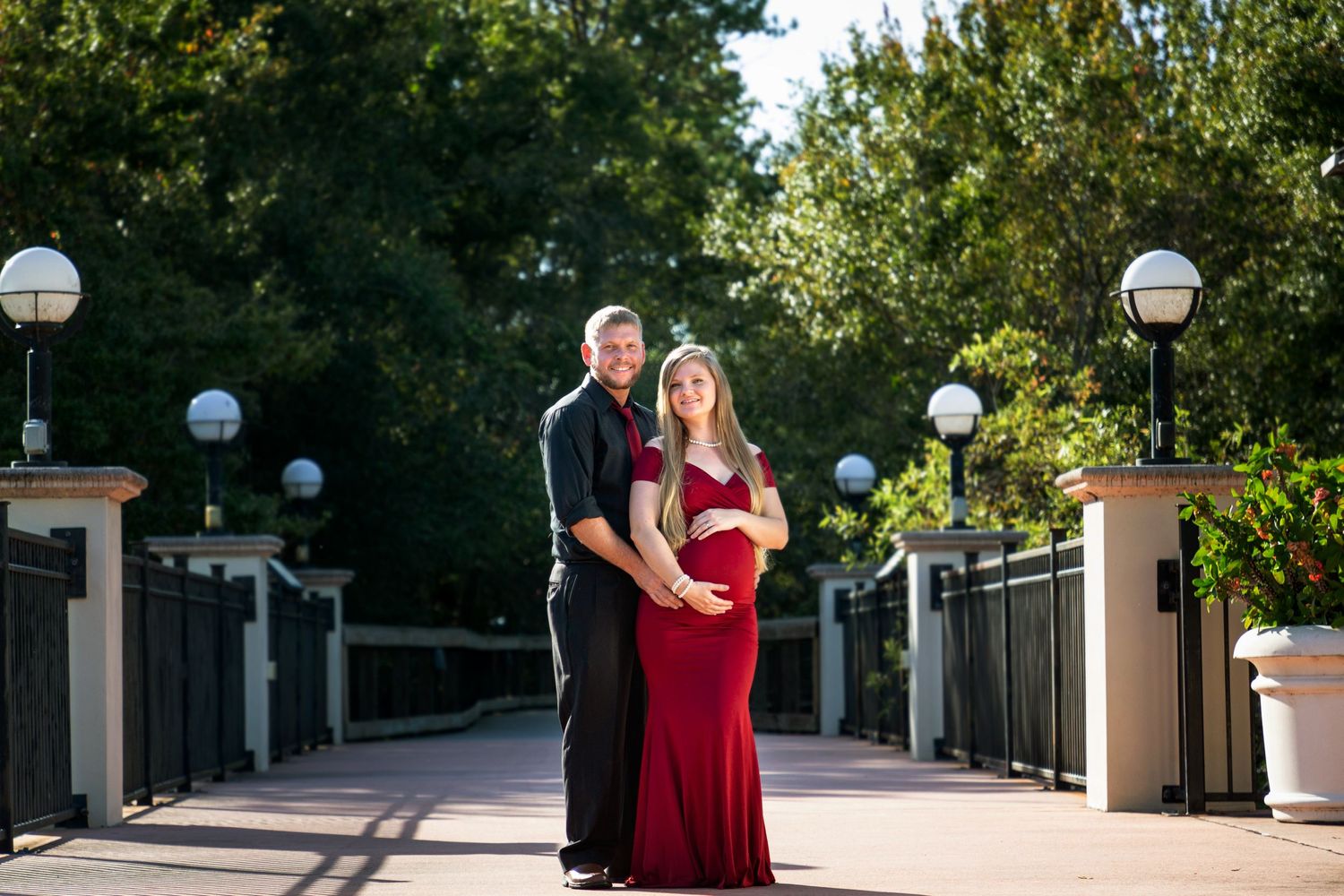 Pregnant woman in red dress standing with man in black shirt and pants holding her pregnant belly while on the bridge.