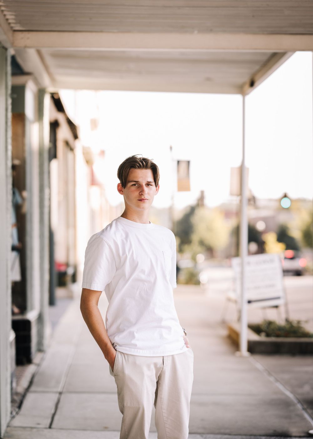 Portrait of someone in a white t-shirt standing under a covered walkway in front of retail stores.
